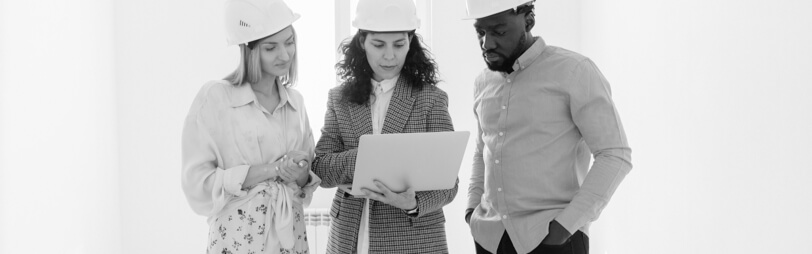 Three individuals in white hats gather around a laptop, discussing plans related to construction and execution.
